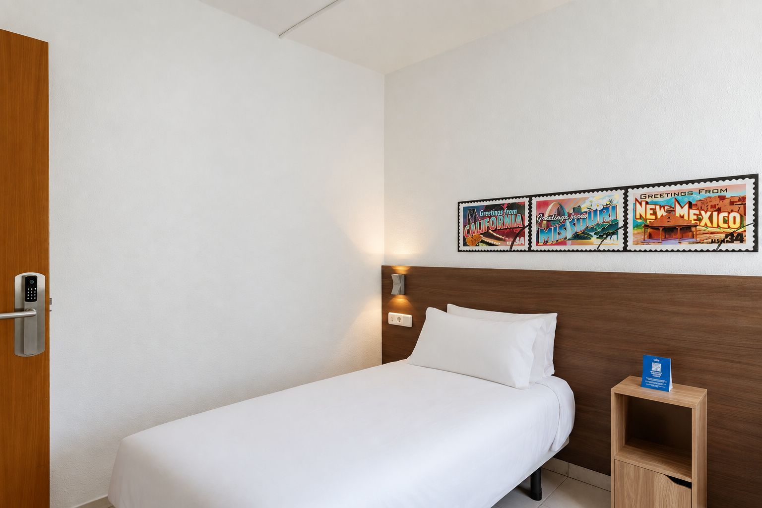 Habitación de hotel con una cama individual, cabecera de madera, mesita de noche con lámpara y un cuadro colgado en la pared con tres postales.