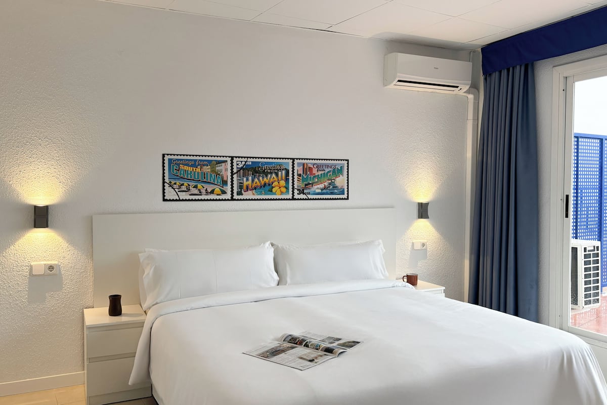 A hotel room with a large bed, two bedside tables, a wall-mounted air conditioner, and a window with curtains.
