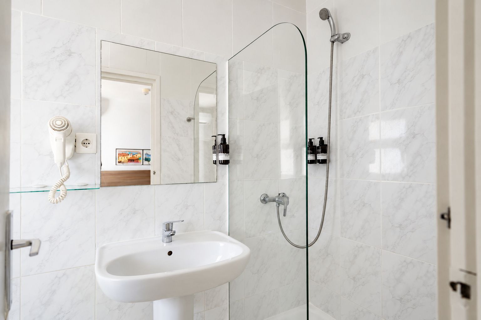 Un baño con lavamanos blanco, cabina de ducha de cristal y espejo con puerta.