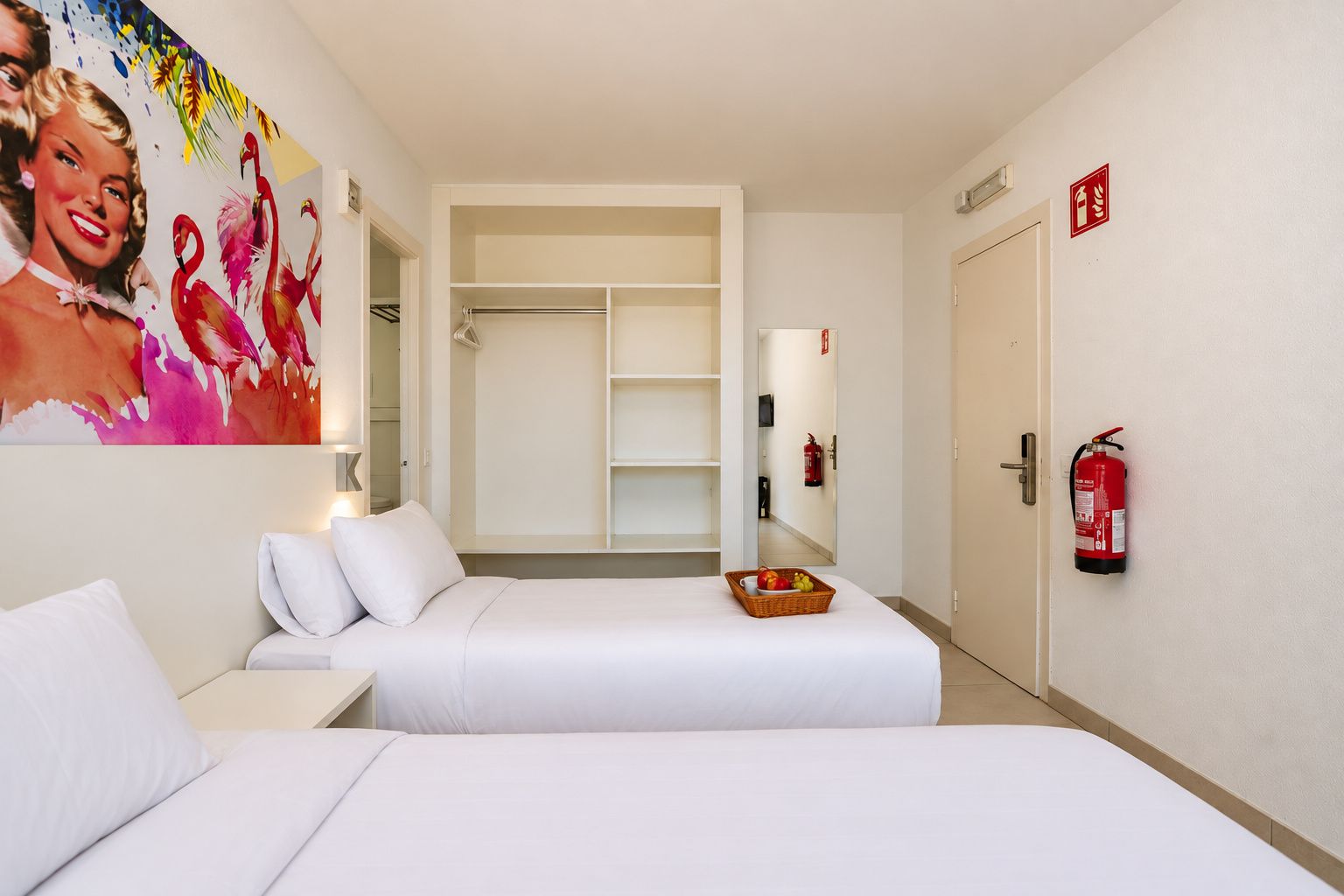 A hotel room with two beds, a wardrobe, a fire extinguisher, and a wall-mounted mirror.