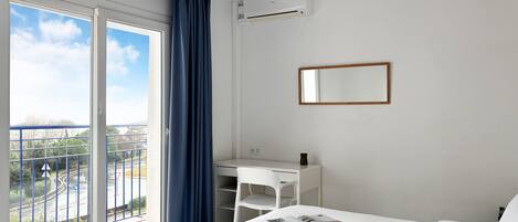 A hotel room with a bed, a desk, a chair, a window with curtains, an air conditioning unit, and a mirror.