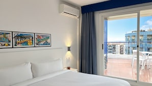 Hotelkamer met een bed, een wandairconditioner, een raam met gordijnen en uitzicht op de stad.