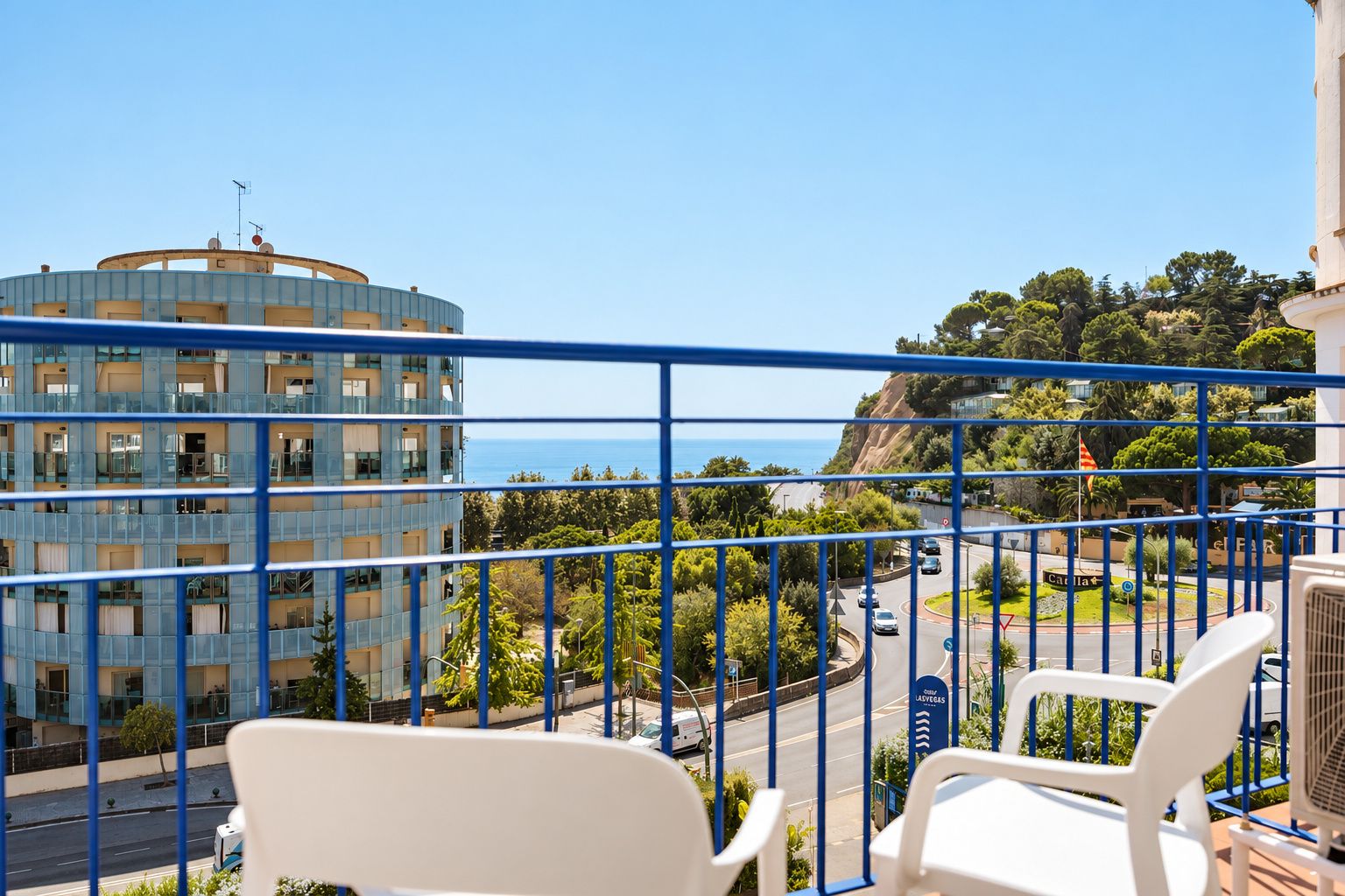 A balcony with a view of a coastal road, a blue building, and a flag.