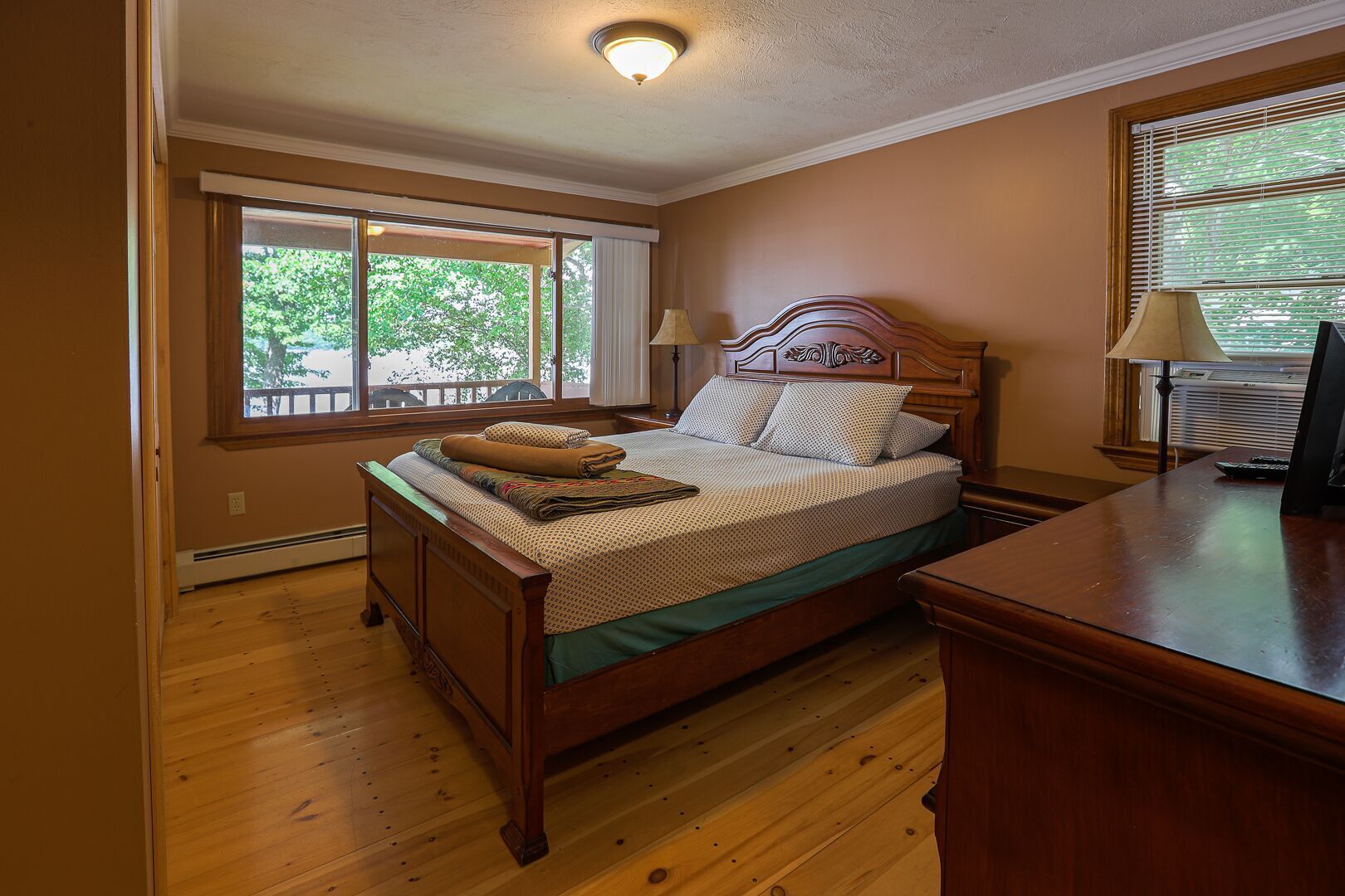 Ein Schlafzimmer mit einem hölzernen Bett, einem Schreibtisch und einem Stuhl. Ein Fenster mit Jalousien und eine Deckenleuchte sind vorhanden.