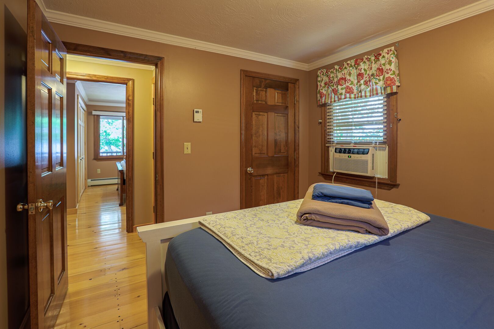 A bedroom with a bed, a window with blinds, a door, and a hallway leading to another room.