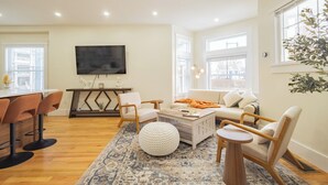 Uma sala de estar moderna com piso de madeira, um sofá branco, uma mesa de centro e uma TV fixada na parede.