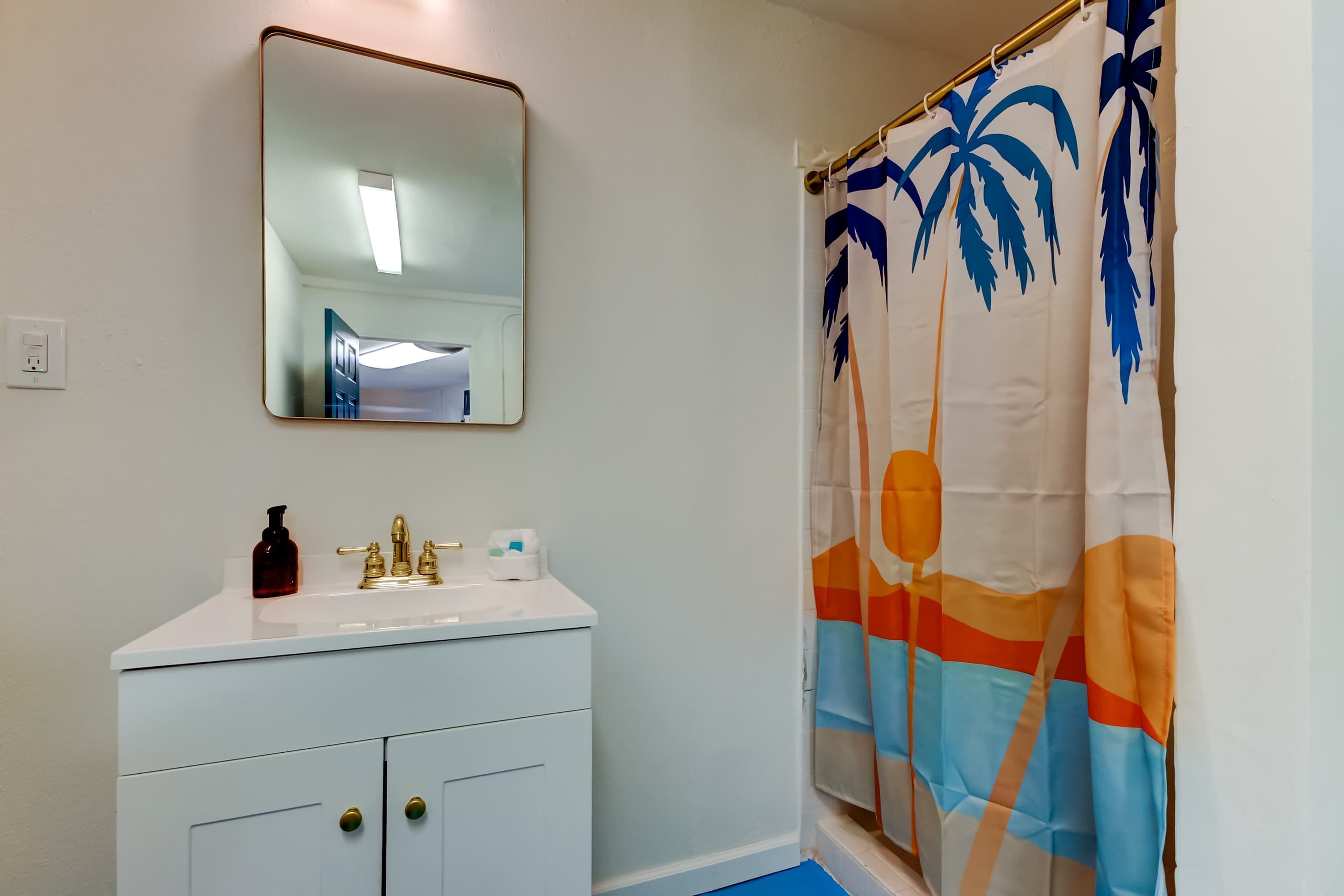 A bathroom with a white sink, gold faucets, a mirror, and a shower curtain with a palm tree design.