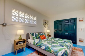 Ein Schlafzimmer mit einem Bett, zwei Nachttischen, einem Fenster mit dekorativer Abschirmung und einer dunkel gefärbten Tür, sowie einem an der Wand befestigten Kunstwerk.