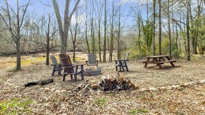 A campsite with a picnic table, two Adirondack chairs, and a fire pit surrounded by trees.