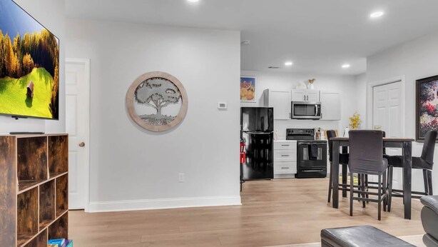 A modern living room with a flat-screen TV, a wooden bookshelf, a dining area with a table and chairs, and a kitchen with white cabinets and appliances.