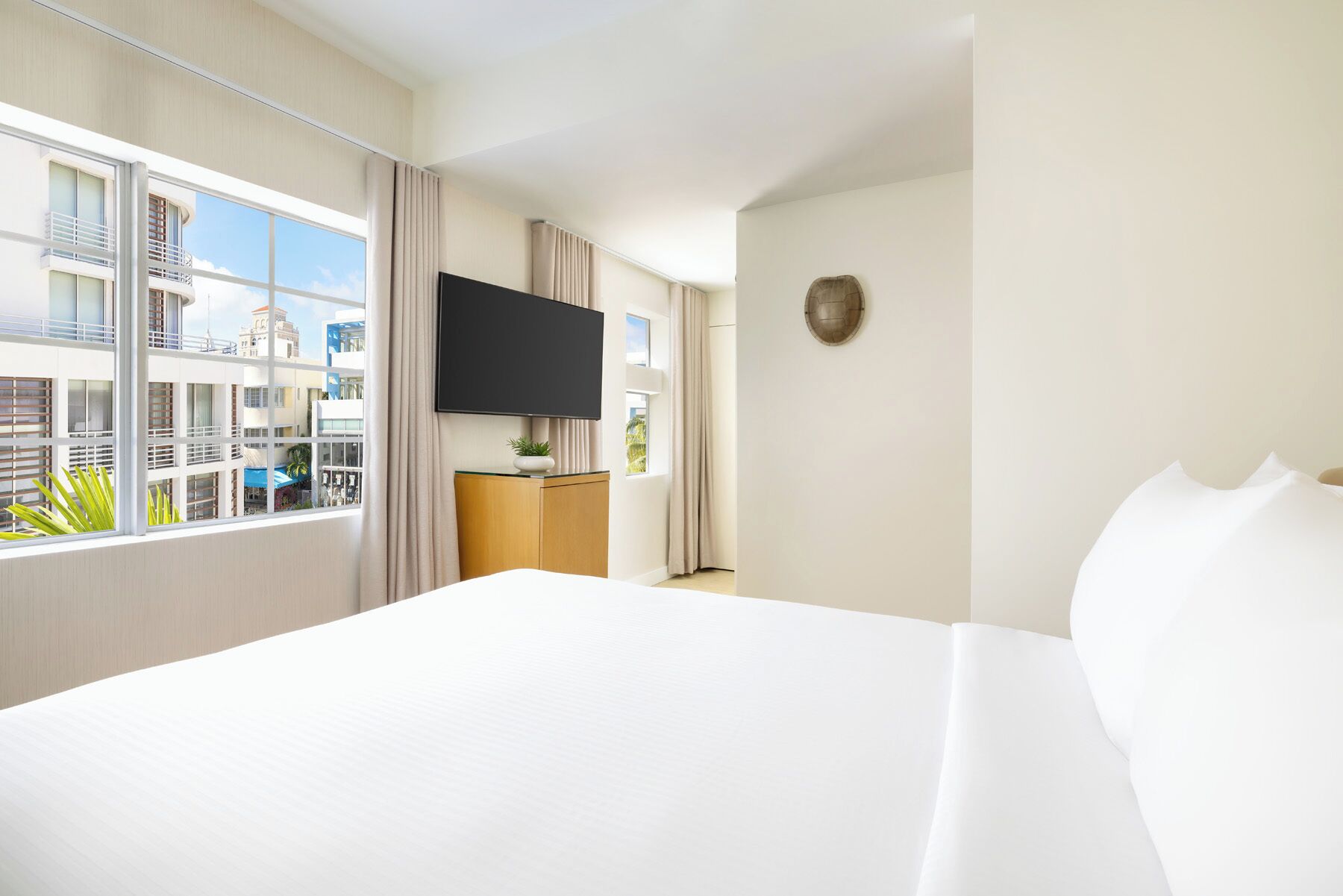 A hotel room with a large bed, a flat-screen TV, a wooden nightstand, and a view of buildings through large windows.