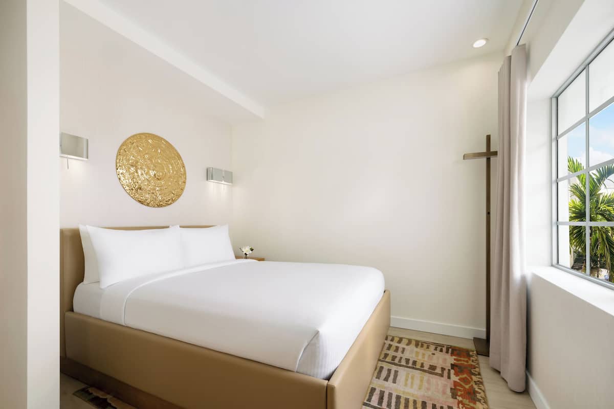 Una habitación de hotel moderna con una cama grande, una lámpara de pared y un ventanal con cortinas.