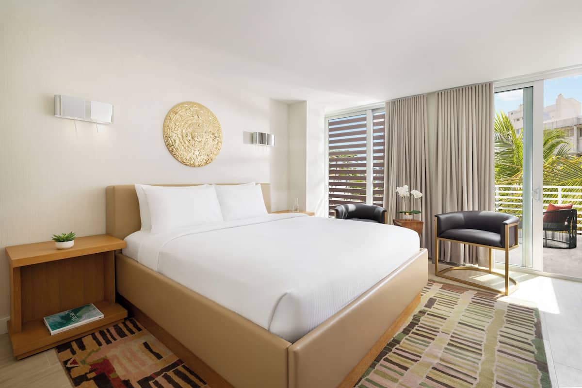 Una habitación de hotel moderna con una cama grande, una mesita de noche con una planta, un adorno en la pared, una silla y vistas al exterior a través de grandes ventanales.