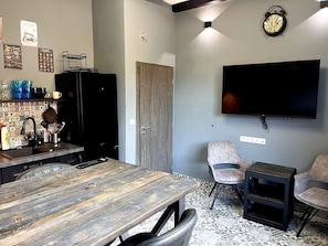 Un salón moderno con una mesa de comedor de madera, dos sillones, una mesita auxiliar, un televisor de pantalla plana montado en la pared y una cocina con refrigerador y salpicadero de azulejos.