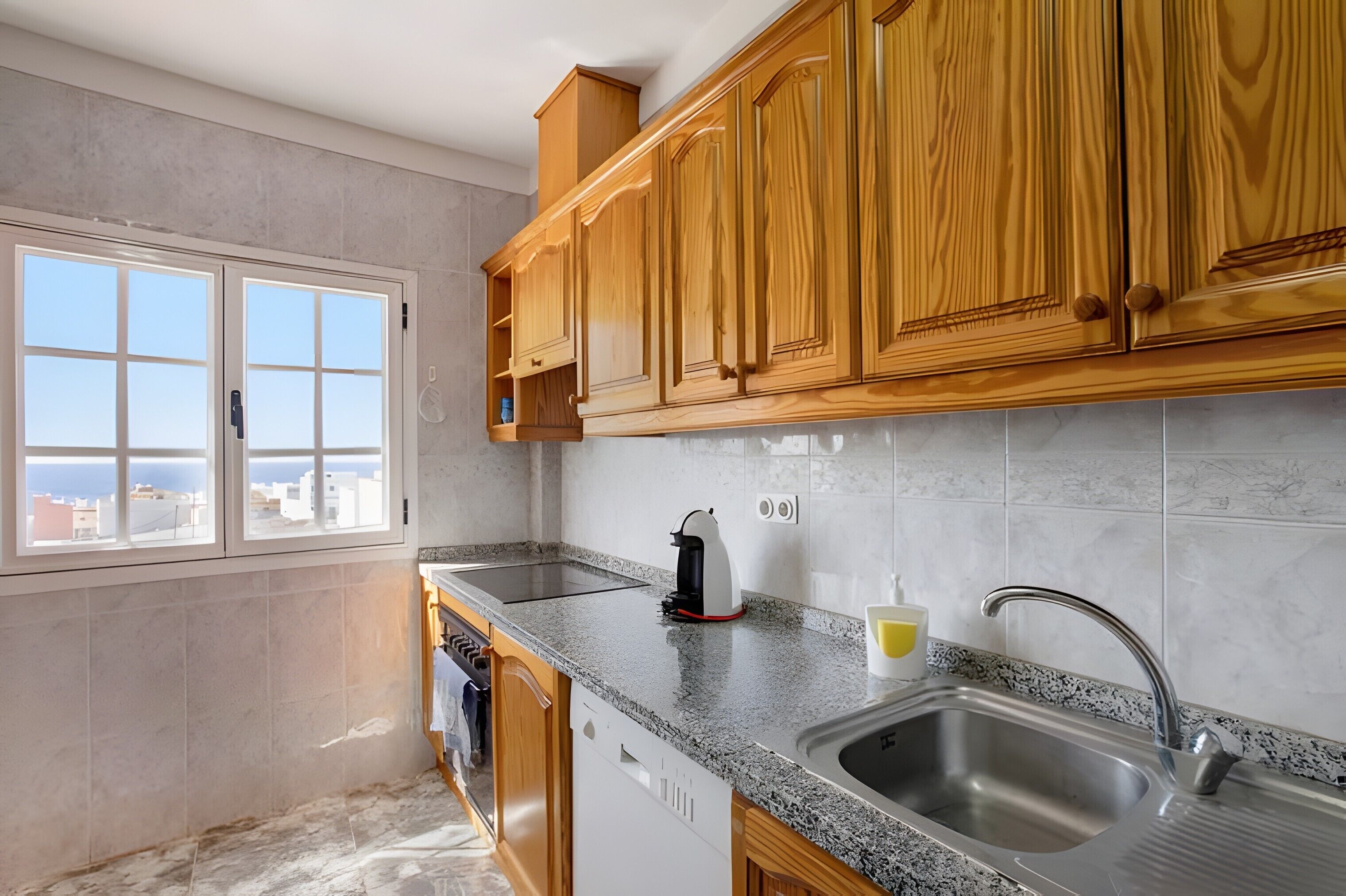 A kitchen with wooden cabinets, a granite countertop, a built-in dishwasher, and a window with a city view.