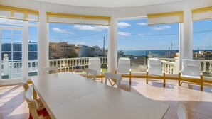 Une pièce dotée d’une grande table et de chaises, de larges fenêtres avec des stores jaunes, et offrant une vue sur la mer.