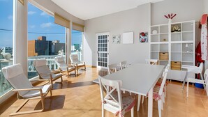Une pièce spacieuse avec une grande table à manger, des chaises blanches, une bibliothèque blanche et une vue sur la ville grâce à de grandes fenêtres.