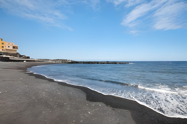 Una playa con una costa curva, un edificio de varios pisos a la izquierda y un cielo despejado.