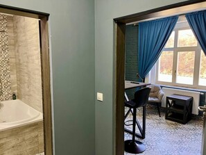 A bathroom with a bathtub and a shower area. There is a window with blue curtains and a view of trees outside. - Contemporary Wood Haven | Center (Karlovy Vary)