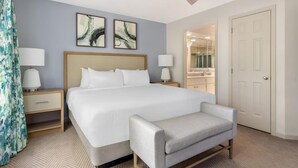 Habitación de hotel con una cama grande, dos mesitas de noche con lámparas, un ventilador de techo, un banco y una puerta que da a un baño.