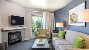 Ein modernes Hotelzimmer mit Flachbildfernseher, Kamin, Sofa mit grünen Kissen, gemustertem Sessel und Esstisch mit Tablett.