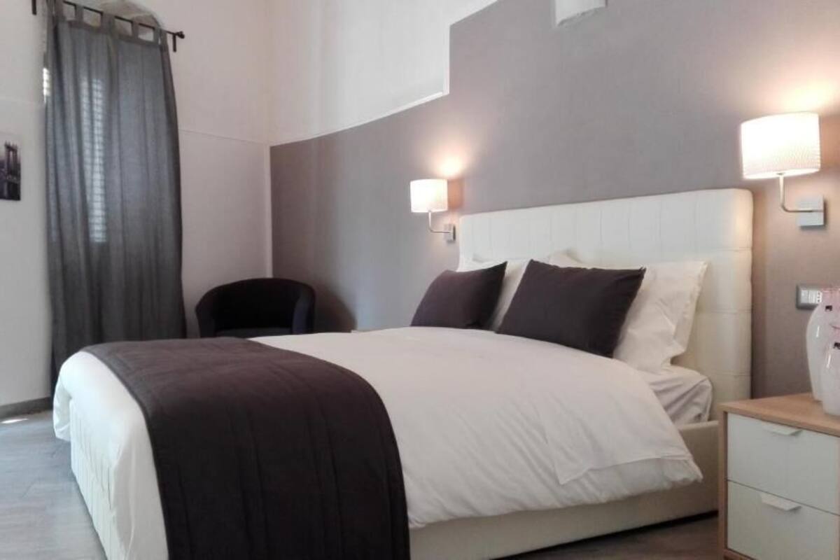 A modern hotel room with a large bed, two bedside lamps, a nightstand, a chair, and a curtain.