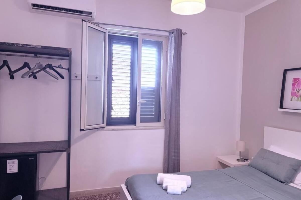 A bedroom with a bed, a desk with a chair, a window with curtains, a wall-mounted air conditioner, and a framed picture on the wall.