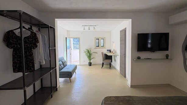 A room with a bed, a TV mounted on the wall, a desk with a chair, a plant, and a clothing rack. - A due Passi Dal Centro (Noto)