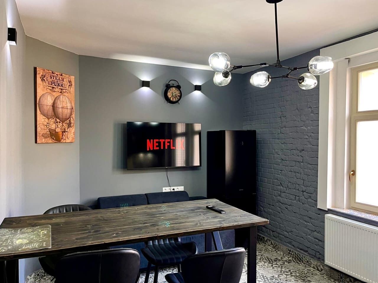 A modern room with a wooden dining table, black leather chairs, a mounted TV displaying 'NETFLIX', and a brick wall.