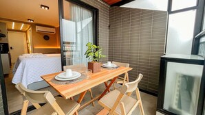 A balcony with a wooden table set for two, a potted plant, and a view of a bedroom with a bed and a TV.