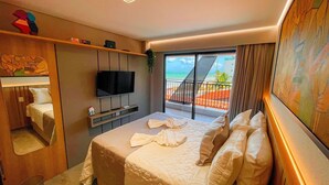 A hotel room with a bed, a TV mounted on the wall, a balcony with a view of the beach, and a small shelf with items on it.