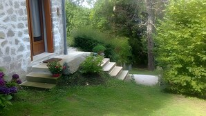 Ein Steingebäude mit einer Holztür, Stufen zu einem Balkon und einem Garten mit verschiedenen Pflanzen.