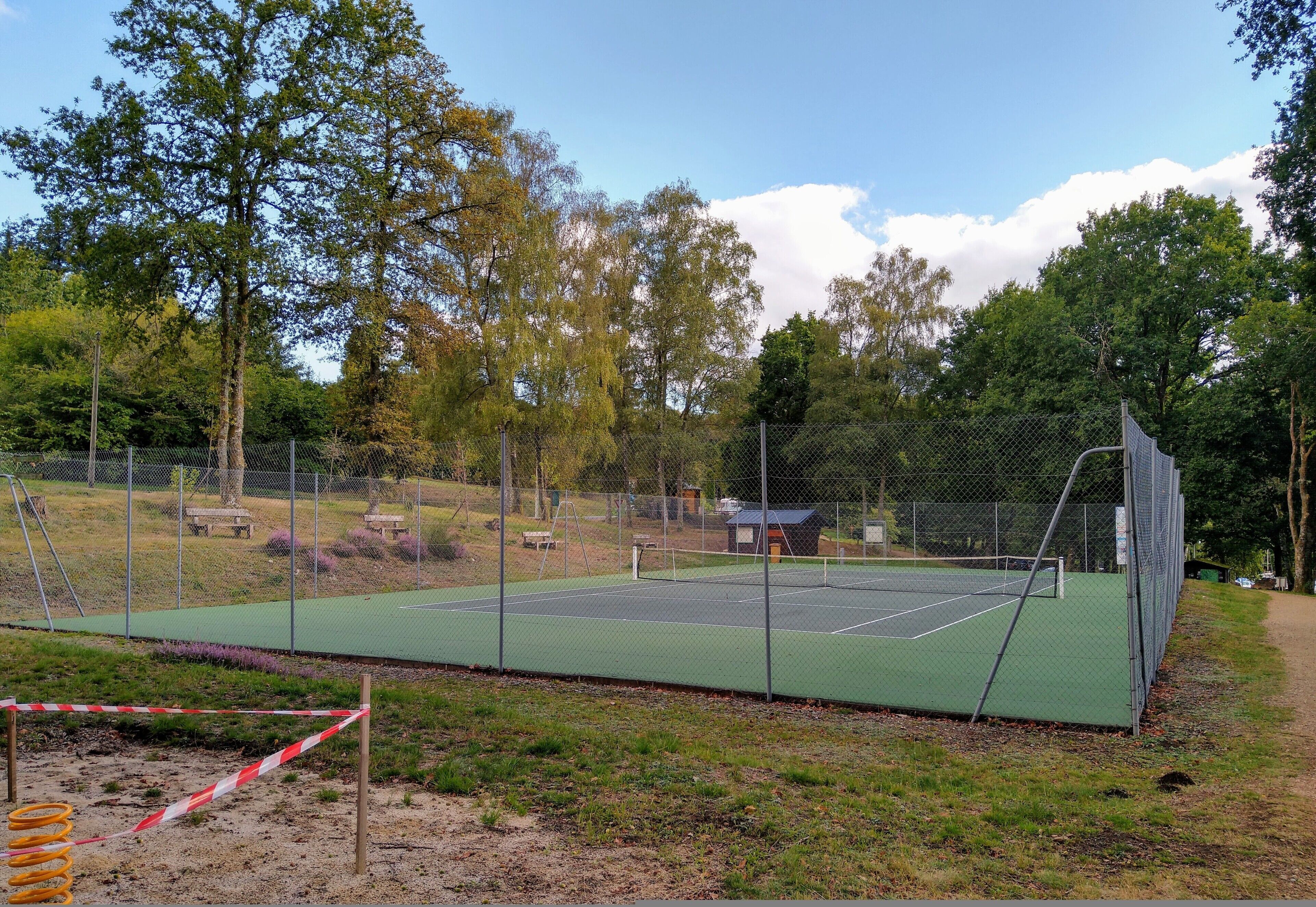 Un court de tennis entouré de clôtures, doté d’une surface verte, et bordé d’arbres et d’un espace pique-nique.