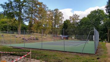 Un court de tennis entouré de clôtures, doté d’une surface verte, et bordé d’arbres et d’un espace pique-nique.