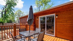 Una terraza de madera con una mesa, sillas, una sombrilla y un ventanal corredizo que da a un espacio exterior.