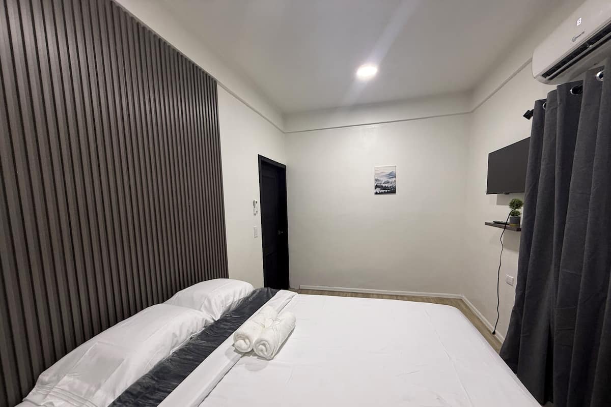 Una cama bien hecha con ropa de cama blanca y gris, un cabecero negro y una toalla enrollada.