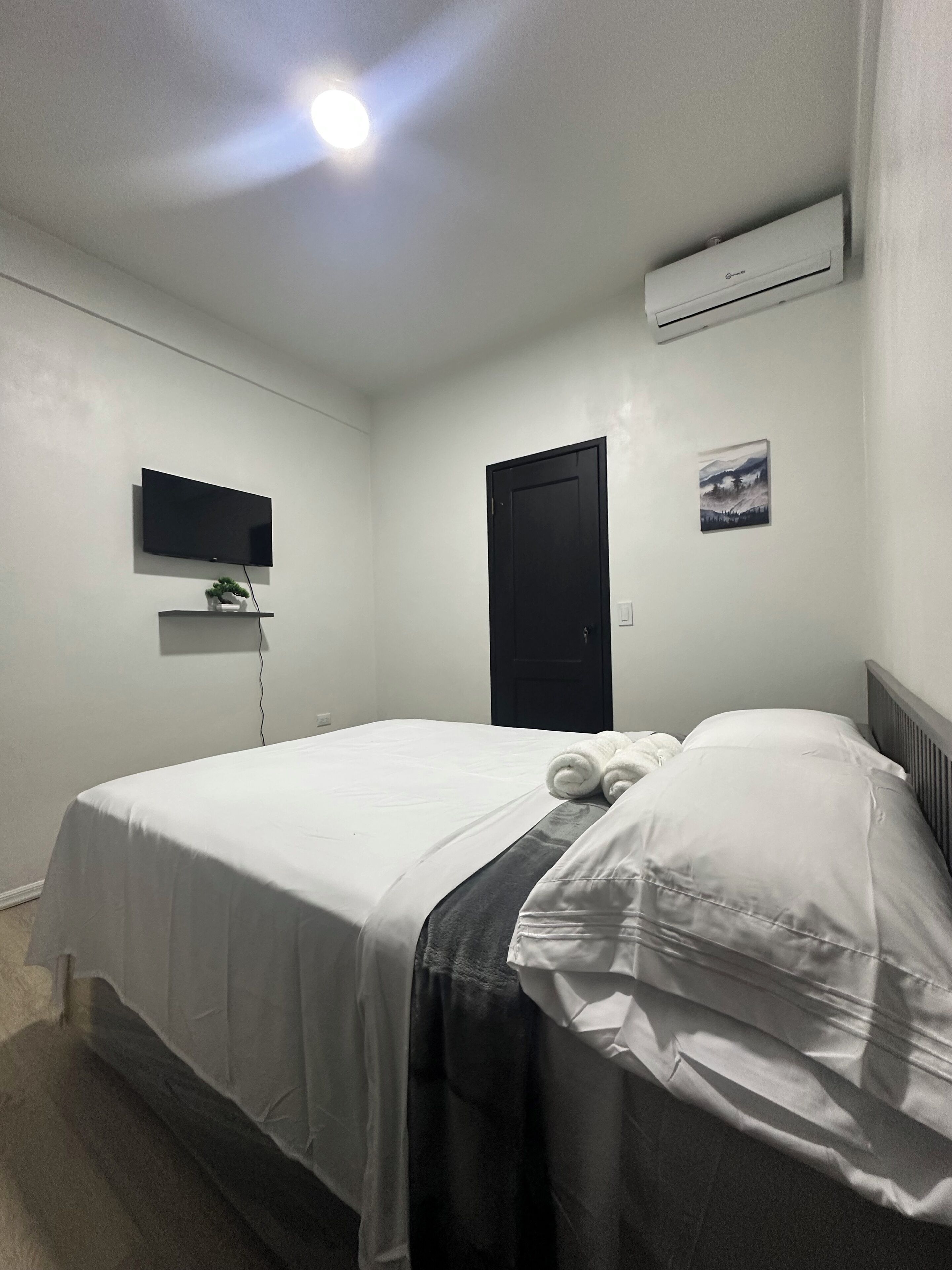 A neatly made bed with white linens, a flat-screen TV mounted on the wall, a black door, and an air conditioning unit.