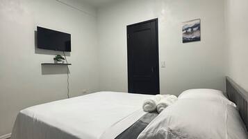 A neatly made bed with white linens, a flat-screen TV mounted on the wall, a black door, and an air conditioning unit.