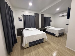 Ett hotellrum med två sängar, mörka gardiner, en vit vägg och ett trägolv.