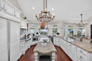 A spacious kitchen with white cabinetry, granite countertops, a central island, and two chandeliers. - 4BR Home | Pool, Hot Tub, Game Room & Fire Pit (Mesa)