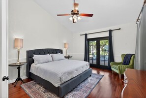 Une chambre à coucher avec un lit, deux tables de chevet, un ventilateur de plafond, une fenêtre avec des rideaux et un fauteuil vert.