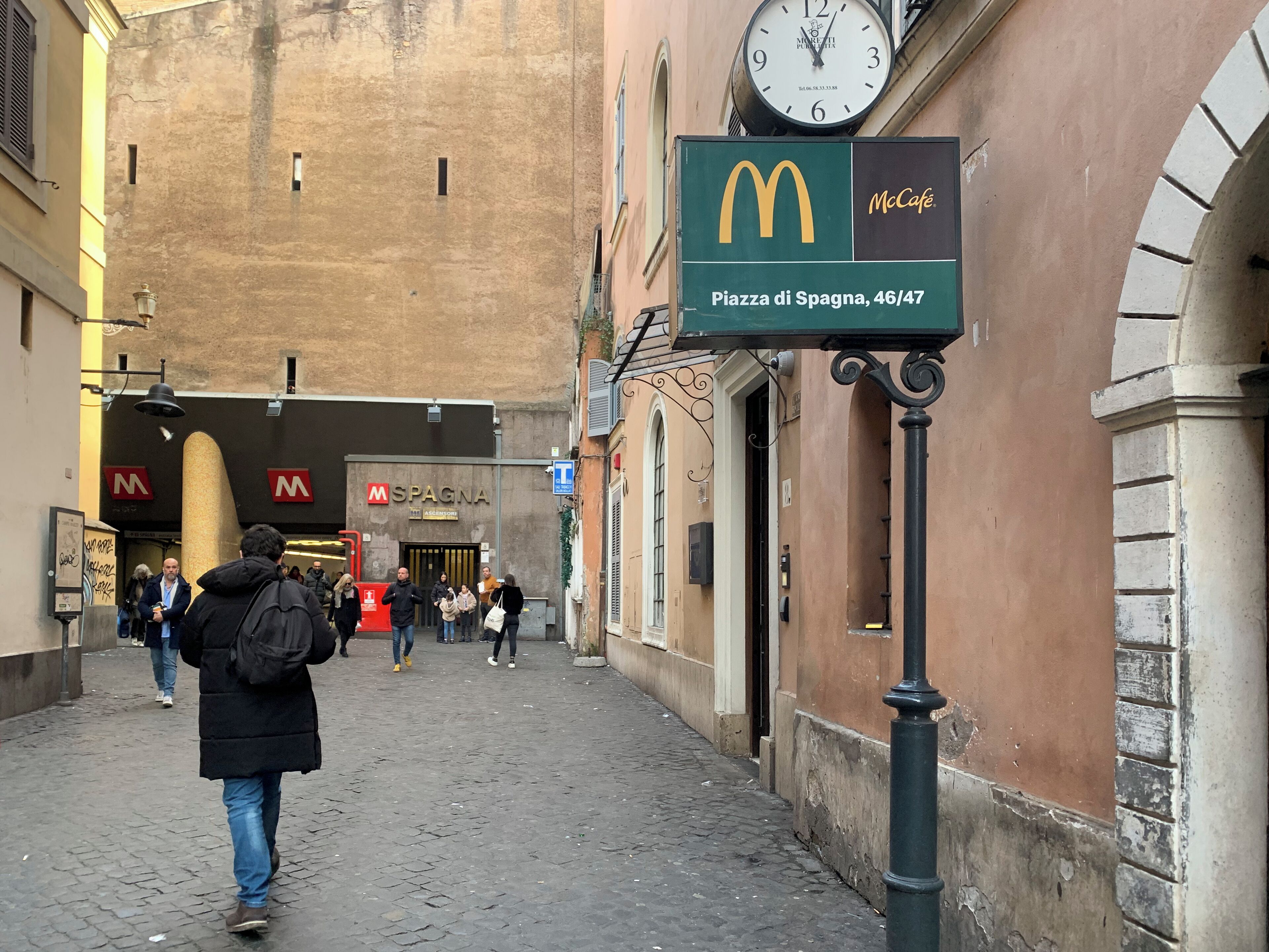 A cobblestone street with a McDonald's sign, a SPAGNA restaurant, and a clock above the entrance.