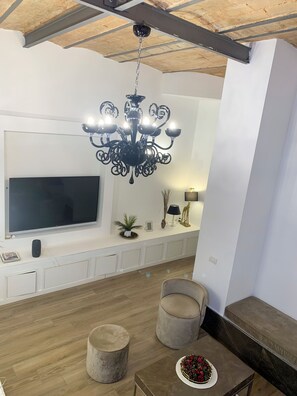 Uma sala de estar moderna com uma grande televisão de tela plana, um candelabro preto, duas poltronas, uma mesa de centro com um arranjo floral e teto de madeira.