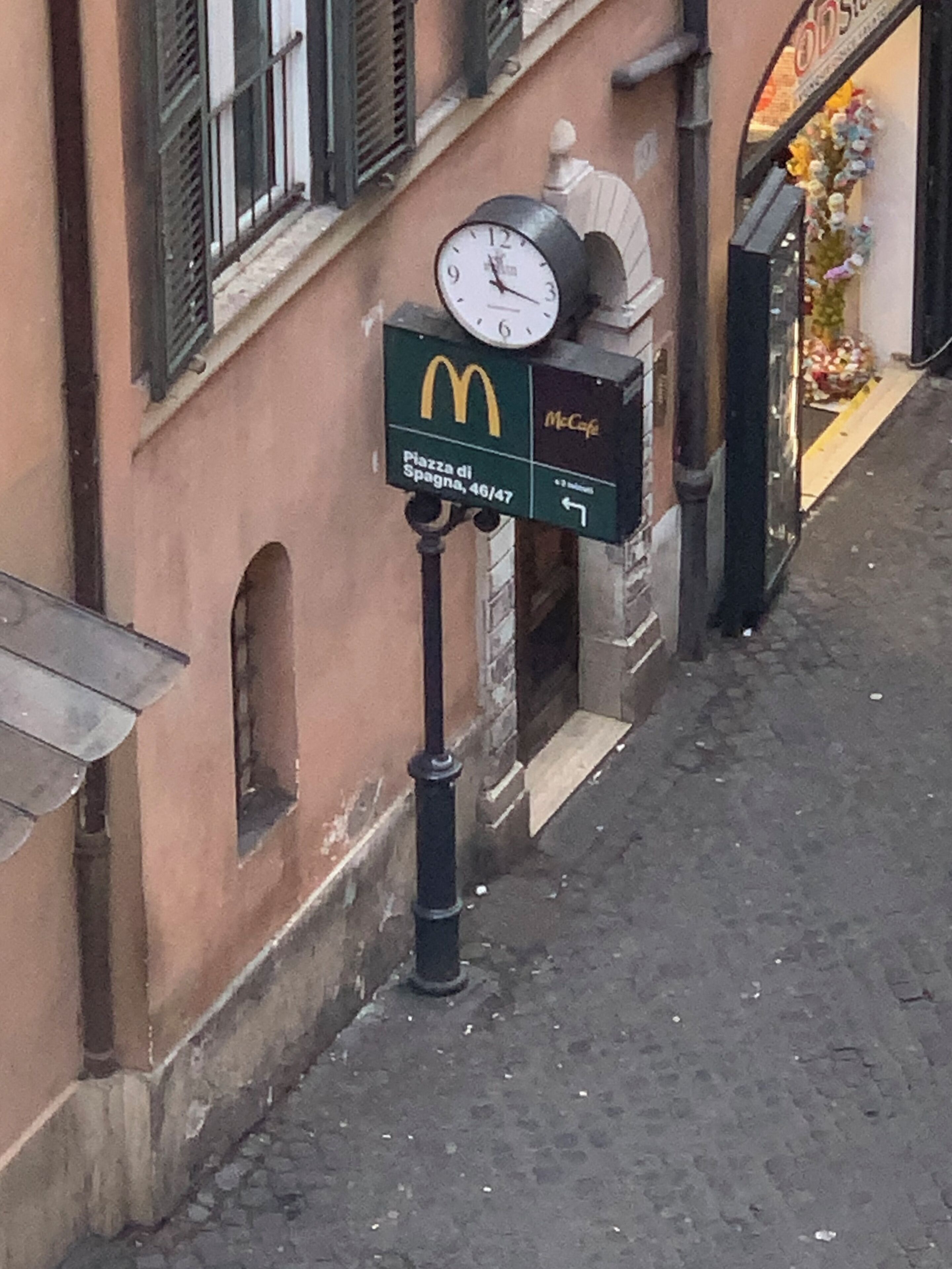 Ein Straßenschild mit einer Uhr und dem McDonald's-Logo, das einen McDonald's-Standort an der Piazza di Spagna 46/47 anzeigt.