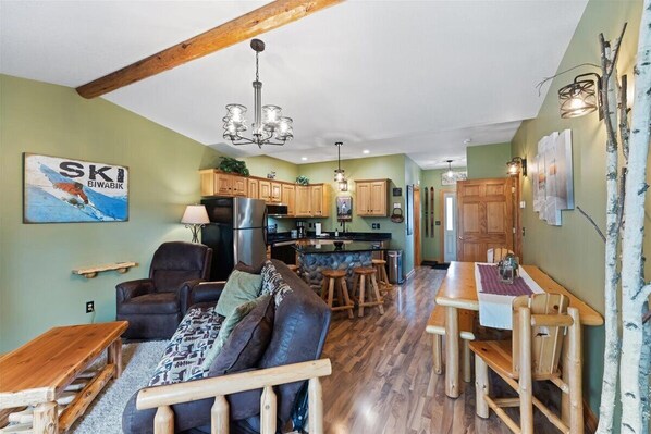 A cozy living area with a sofa, a wooden coffee table, and a dining table with chairs. A kitchen with wooden cabinets and a refrigerator is visible in the background.