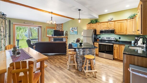 A modern kitchen with wooden cabinets, a central island with stools, a built-in oven, and a large window overlooking a garden.