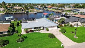 A residential area with a house featuring a large garage, a well-maintained lawn, and a dock extending into a body of water. - Sunset Reach ...Waterfront Family Pool Home - Gulf Access -  Sunset Views - (Cape Coral)