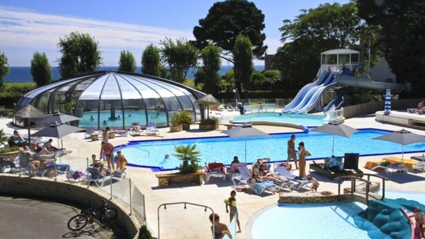 A pool area with a covered section, a water slide, and lounge chairs. - ZEcamping 4 étoiles 200m plage piscine couverte locatif TV LV BBQ 35m2 (Névez)