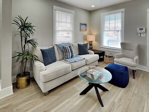 En stue med en beige sofa, et glasbord, en blå skammel, en potteplante og en lænestol.