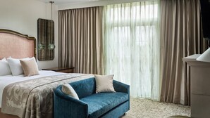 Camera d'albergo con un letto grande, una poltrona blu, una testiera in legno, un comodino, uno specchio e un'ampia finestra con tende.